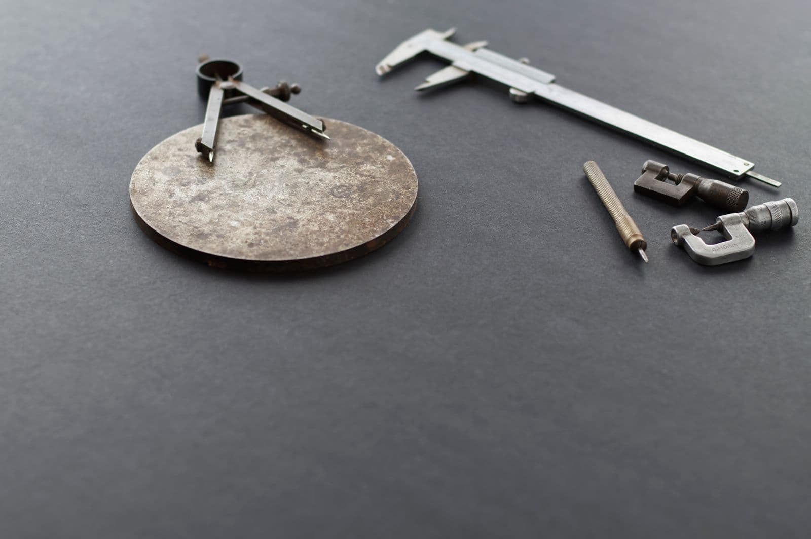 An undocumented machined steel disc next to a vernier caliper, micrometer and divider compass on a dark surface